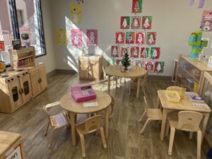 Pre-K room at Howard Burnham, showing appropriately-sized chairs and tables, a kitchen play area, art supplies, and student art on the wall.