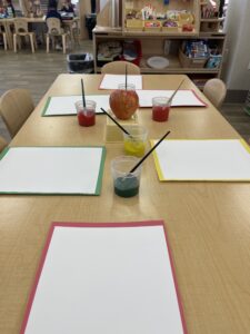 Red, yellow, and green paint in cups on a table. The table also has several blank pieces of paper and an apple elevated on a plastic container. More art supplies are visible in the background.